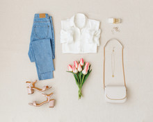 Blue Jeans, White Shirt, Heeled Sandals,  Small Cross Body Bag With Chain Strap, Pink Tulips On Beige Background. Woman's Stylish Spring Summer Outfit. Trendy Women's Clothes. Flat Lay, Top View.