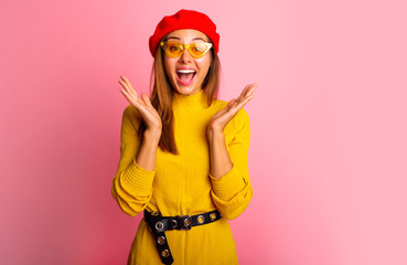 Wall Mural - Surprised young woman in yellow dress, red hat and yellow glasses over gpink background.Happy surprised young lady laughing and holding her hands near cheeks