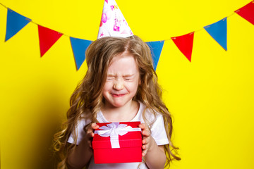 Happy child received a birthday present. Birthday girl with a gift smiles closing her eyes on a yellow background. Red gift box in children's hands.