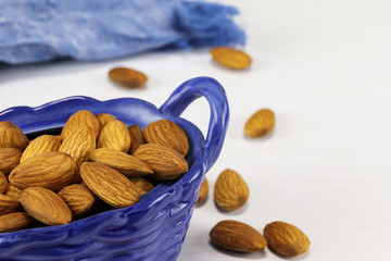 Almond nuts lie in a blue glass basket and on a white surface