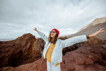 Portrait Of A Carefree Stylish Woman Dressed In Jeans And Red Hat Enjoying Trip On A Rocky Ocean Shore Near The Lighthouse, Traveling On North-west Of Tenerife Island, Spain