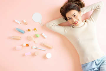 Pretty Asian woman smiling while lying on pink floor with her cosmetic makeup tools. Top view