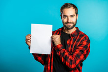 Handsome Man Holding Vertical White A4 Paper Poster. Copy Space. Smiling Hipster Guy In Red Plaid Shirt On Blue Background.