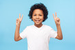 © khosrork - Portrait of happy and victorious little boy in T-shirt showing victory or peace gesture and smiling, doing v sign, number two, cute child enjoying win. indoor studio shot isolated on blue background