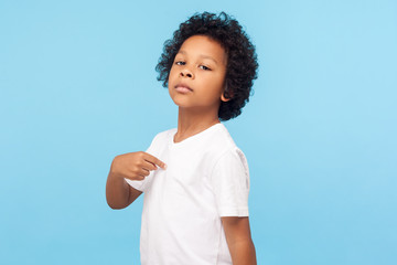 This is me. Portrait of preschool curly boy in T-shirt being proud and arrogant pointing at himself with serious supercilious expression, egoistic child. indoor studio shot isolated on blue background