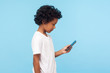 Side view of serious nice preschool boy with curly hair in T-shirt attentively reading message on smartphone, using mobile application, social media. indoor studio shot isolated on blue background