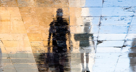 Blurry reflection shadow silhouettes of a man and a boy walking on a wet street on a sunny summer day