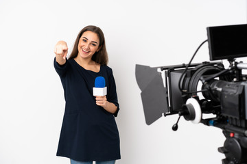 Reporter woman holding a microphone and reporting news isolated on white background points finger at you while smiling