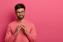 Handsome Unshaven Man Has Tricky Evil Plan, Steepls Fingers And Looks Mysteriously At Camera, Thinks Over Good Idea, Wears Casual Sweater And Spectacles, Poses Over Rosy Background, Copy Space