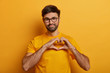 © WHstudio Leushin N - Pleased good looking man with thick bristle, shapes heart over chest, confesses in love, has romantic mood, expresses truthful feelings, wears yellow t shirt. My heart completely belongs to you