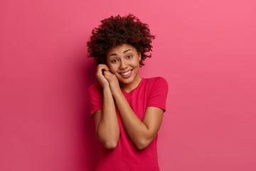 Wall Mural - Tender pleasant looking woman keeps hands near face, has positive happy look, dressed in casual red t shirt, concentrated on pleasant thoughts, isolated on pink background, rejoices spare time