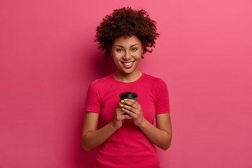Wall Mural - Pleasant looking smiling ethnic woman enjoys freshly brewed coffee, holds paper cup, has energy drink to feel refreshed in morning, wears casual t shirt, has happy expression, models in studio