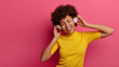 © WHstudio Leushin N - Overjoyed African American woman listens modern music via wireless headphones, enjoys good sound and chills with eyes closed, smiles broadly, wears casual yellow t shirt, isolated on pink wall