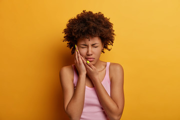 Wall Mural - Displeased dark skinned woman suffers from toothache, touches cheek, needs to see dentist, take painkillers, has health problems, closes eyes with pain, dressed casually, isolated on yellow background