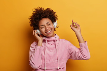 Poster - Happy female teenager enjoys pleasant music, wears stereo headphones, dances with arm raised, feels upbeat, catches every tune of song, wears rosy velvet sweatshirt, isolated on yellow background