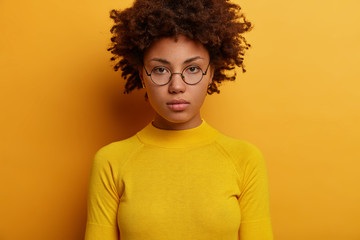 Wall Mural - Headshot of serious looking woman with Afro hairstyle, looks directly at camera, wears round spectacles and yellow jumper, thinks over about something important, stands in studio. Face expression