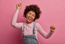 Happy Dark Skinned Girl Enjoys Every Moment Of Life, Dances And Moves, Raises Arms And Clenches Fists, Closes Eyes, Has Good Mood, Wears Denim Sarafan And Turtleneck, Isolated On Pink Background