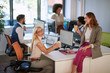 © luckybusiness - two female colleagues talking, drinking coffee and smiling at work, with group of colleagues in the background. modern, office, casual,  business concept