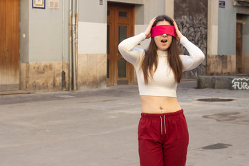 Portrait of attractive caucasian young woman with eye tape being surprised, opening her eyes and mouth with shock, touching her head with hands, in the city, white top and red pants, dark air. Place f