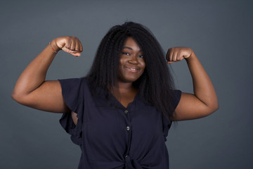 Wall Mural - Waist up shot of African woman raises arms to show her muscles feels confident in victory, looks strong and independent, smiles positively at camera, stands against gray background. Sport concept.