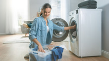 Beautiful And Happy Brunette Young Woman Comes Towards The Washing Machine In Homely Jeans Clothes. She Loads The Washer With Dirty Laundry. Bright And Spacious Living Room With Modern Interior.