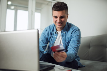 Smiling young handsome man using credit card and laptop at home