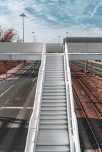A Vertical View Of A White Long Metal Stairway To An Overhead Passage For Pedestrians With Railway Tracks Below On The Right And An Urban Road On The Left, Sunny Day, Lisbon, Portugal