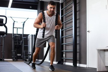 Athletic young man with battle rope doing exercise in functional training fitness gym.