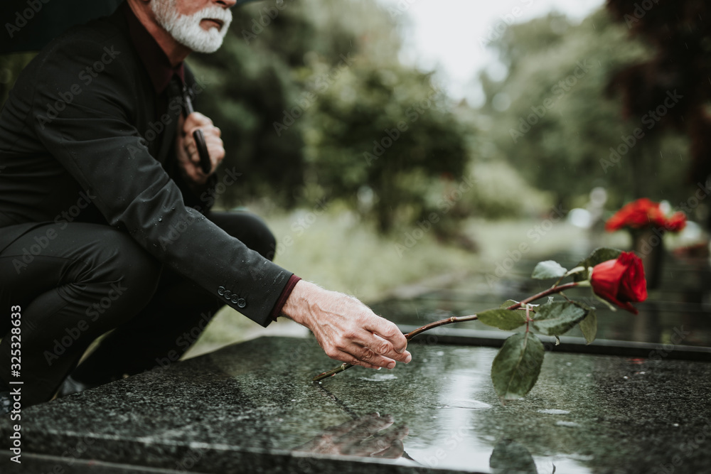Elegant sad elderly man standing on the rain with umbrella and grieves at the grave of a loved person