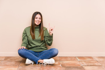 Wall Mural - Young caucasian woman sitting on the floor isolated showing a horns gesture as a revolution concept.