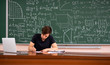 © Mihail - young male teacher sitting at the table near blackboard and check the exam works of pupils