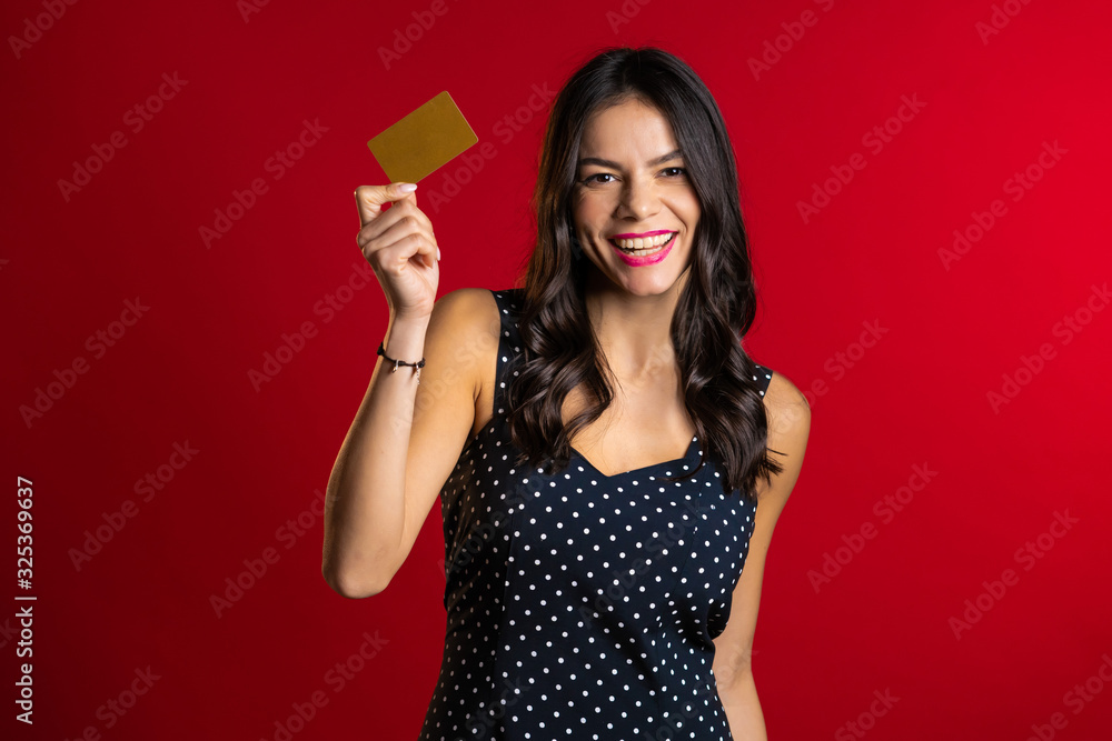 Successful pretty latin girl look to camera and showing unlimited gold credit card on red studio background.  - obrazy, fototapety, plakaty 