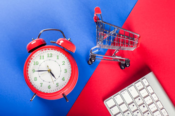 Alarm clock, shopping trolley and keyboard on the doubble color background. Online shopping and sales concept