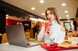 © Тарас Нагирняк - Blonde and happy woman in white sweater student is learning with a laptop in a cafe, eating fast food and drinking a coca cola.