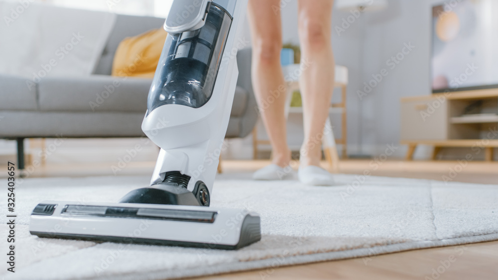 Close Up Shot of a Young Beautiful Woman in Jeans Shirt and Shorts Vacuum Cleaning a Carpet in a Bright Cozy Room at Home. She Uses a Modern Cordless Vacuum. She's Happy and Cheerful.