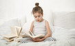 © puhimec - Cute little girl reading a book on the bed in the bedroom.