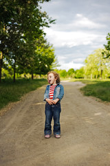 Wall Mural - Happy, funny, little girl having fun outdoors. Adorable, cute curly child, kid fooling, making funny faces, grimaces and showing tongue on the road beautiful summer day.