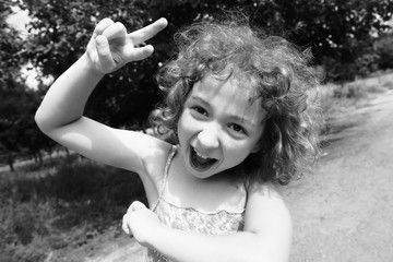 Wall Mural - Happy, funny, crazy little girl having fun outdoors in the summer. Adorable, cute curly child, kid fooling, making funny faces, grimaces on beautiful summer day. Black and white photo.