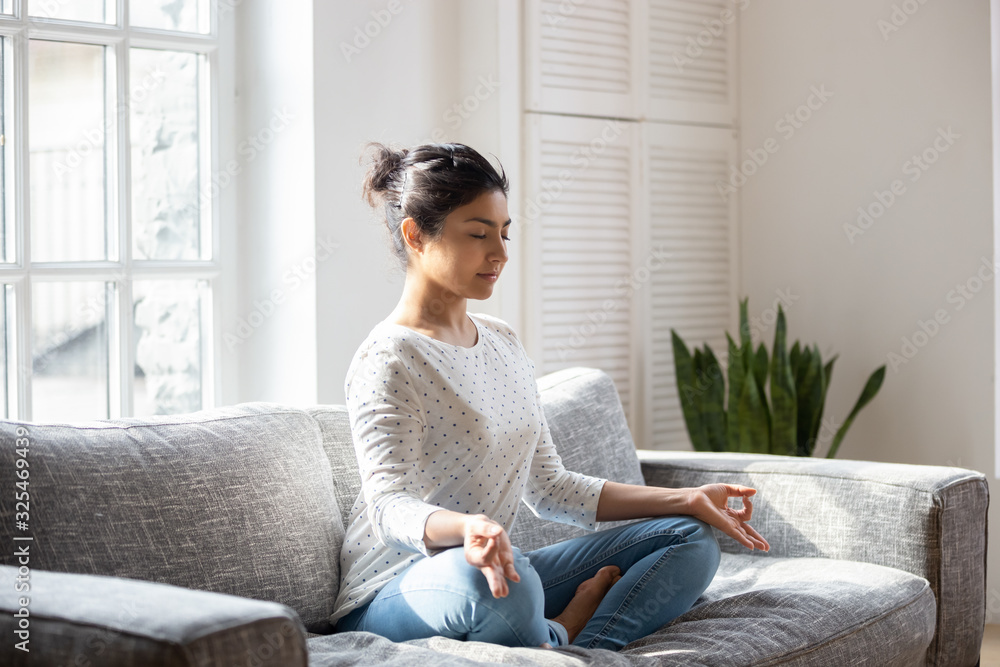 Indian female sitting on couch lotus pose do yoga exercise - obrazy, fototapety, plakaty 