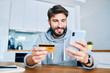 © baranq - Young man sitting at home paying for food online with smartphone and credit card