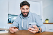 © baranq - Smiling young man sitting at home and paying online with credit card and smartphone