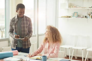 Wall Mural - Portrait of cheerful African-American student looking at curly haired girl while discussing creative project in modern white office, copy space