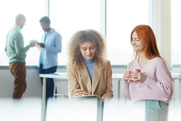 Wall Mural - Portrait of two colorful young women discussing work project during coffee break in office, young intern presenting ideas to manager, copy space