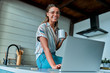 © Valerii Apetroaiei - Enjoying time at home. Beautiful young smiling woman working on laptop and drinking coffee while sitting on the countertop in the kitchen at home.