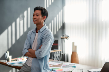 Confident designer. Side view of a young asian man in casual wear with crossed arms looking away and smiling while standing in the modern office