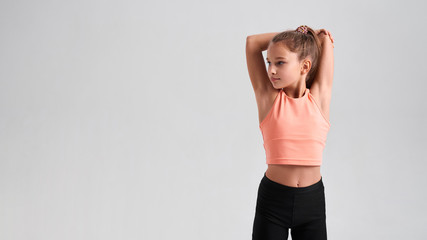 Stay healthy. Flexible cute little girl child looking aside while stretching her body isolated on a grey background. Sport, training, fitness, active lifestyle concept. Horizontal shot.