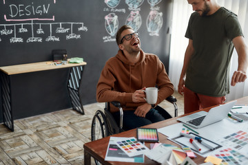 Great mood for work. Young and happy male UX UI designer in a wheelchair holding cup of coffee and laughing while discussing some ideas with his colleague in the creative office
