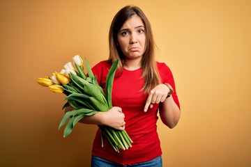 Poster - Young blonde woman holding romantic bouquet of tulips flowers over yellow background Pointing down looking sad and upset, indicating direction with fingers, unhappy and depressed.