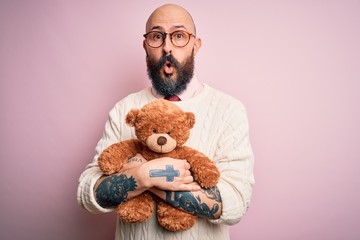 Sticker - Handsome bald man with beard and tattoo holding teddy bear over isolated pink background scared in shock with a surprise face, afraid and excited with fear expression