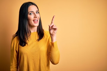 Young brunette woman with blue eyes wearing casual sweater over yellow background showing and pointing up with finger number one while smiling confident and happy.